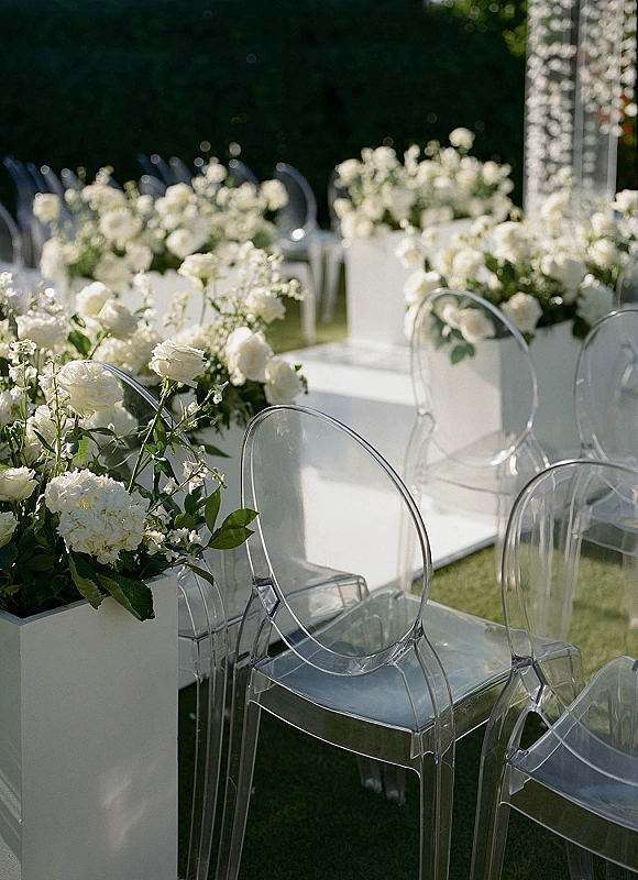 Ceremony aisle design with outdoor ceremony aisle lined by white rose and hydrangea planter boxes, clear acrylic chairs, and a white runner on grass lawn