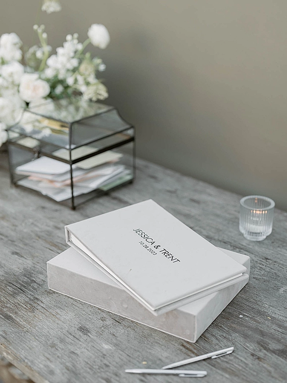 Wedding guest book with a white cover in a presentation box beside pen and ribbed glass votive candle on a weathered wood table