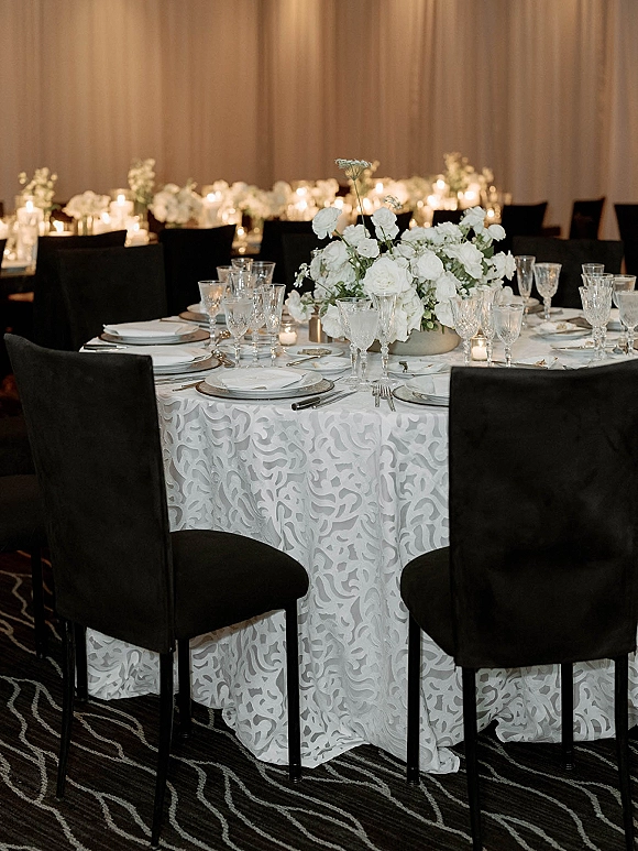 Reception tablescape with a round wedding table setup, white floral centerpiece and votive candles, silver chargers, crystal stemware in dim ballroom lighting