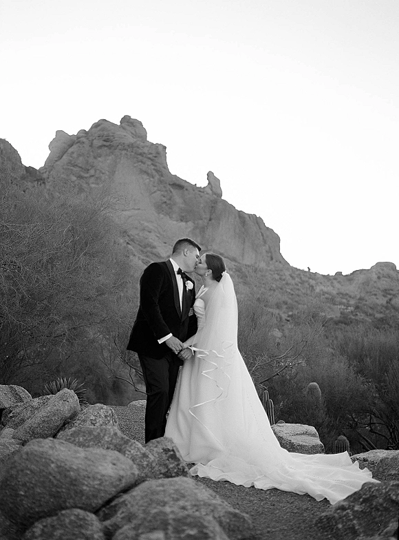 Wedding kiss portrait of bride and groom kissing, her long veil blowing, against desert mountains with boulders, cacti, and open sky