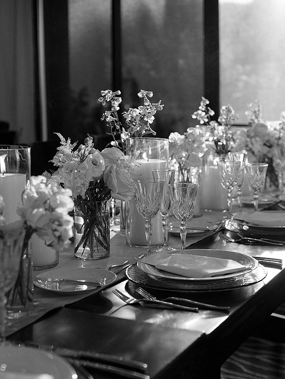 Reception tablescape wedding table setting with glass cylinder candles, white floral bud vases, and crystal stemware on a sunlit banquet table.