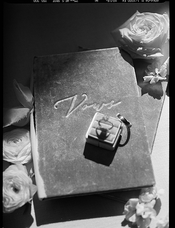 Wedding vows book with rings beside a ring box, ribbon, and white roses on linen fabric, lit by soft sunlight shadows