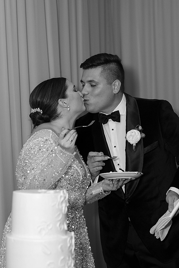 Wedding cake cutting as bride and groom kiss, holding plates and forks beside a white tiered cake against draped curtains
