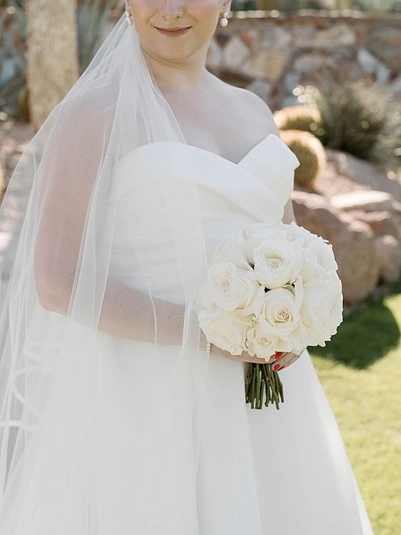Bridal portrait of a bride in a strapless wedding dress with tulle veil, holding a white rose bouquet by a stone wall in a garden