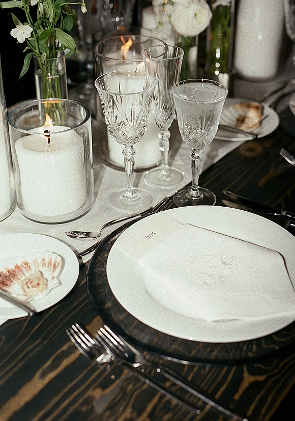 Reception tablescape with black and white place setting, calligraphy place card on white plate and black charger, crystal glasses on dark wood table