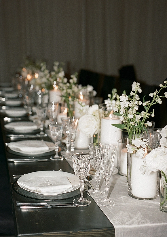 Reception tablescape with white florals, pillar and votive candles, crystal glassware, and white plates set along a black table runner by draped backdrop