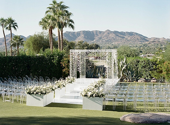 Outdoor ceremony setup with clear acrylic chairs flanking a white aisle, leading to a platform stage beneath hanging white florals, palm trees and mountains beyond
