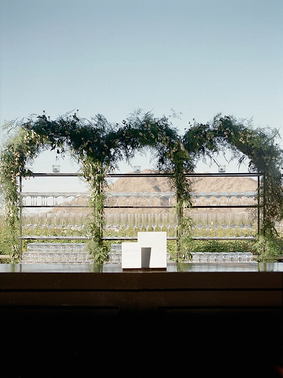 Wedding bar setup with hanging greenery accent, bar shelving and champagne flutes on a black counter against mountains and open sky