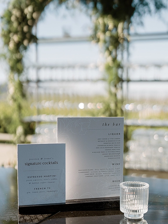 Wedding bar menu and signature cocktails sign displayed on a tabletop with a glass candle holder, set against an outdoor ceremony arch and white chairs