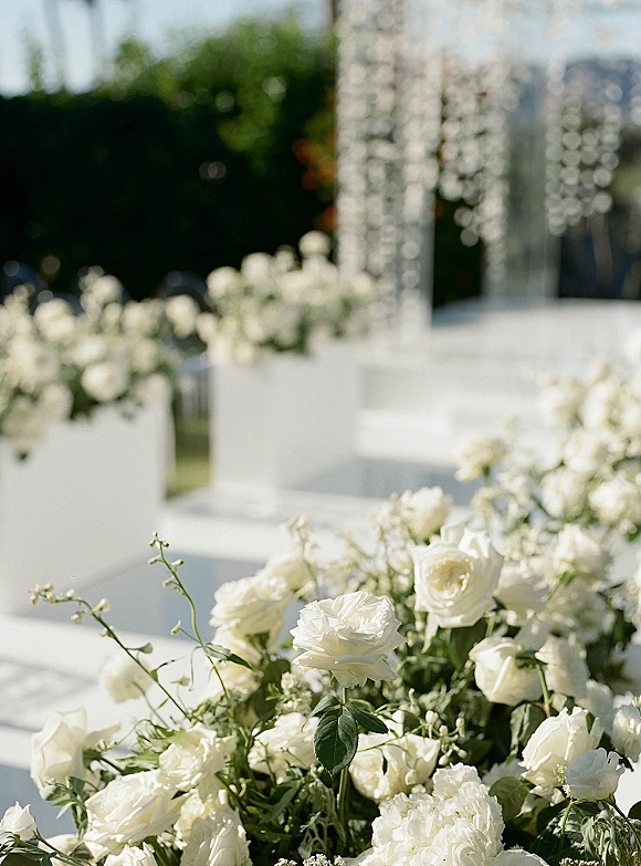 Ceremony aisle decor with white rose aisle flowers in pedestal planters lining a white platform, set against greenery hedges and reflective columns