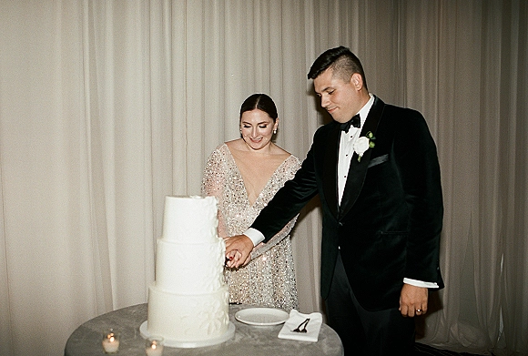 Wedding cake cutting as bride in a sequin dress and groom in tuxedo slice a three-tier cake on a candlelit dessert table before draped curtains