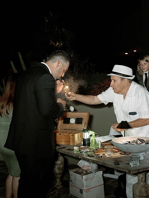 Cigar rolling station wedding cigar bar with cigars, cutter, ashtray, and whiskey tumblers on a lit table in a nighttime garden setting