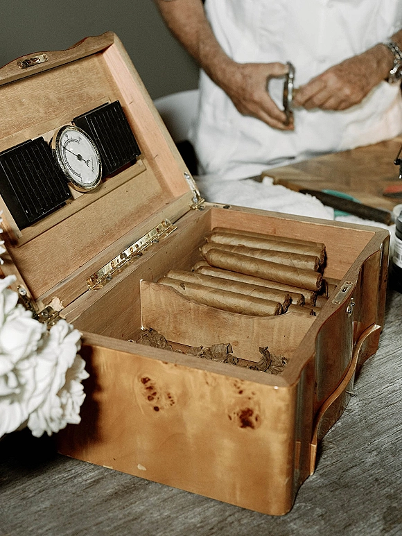 Cigar bar detail with wood cigar humidor, cigars, and cutter on a tabletop, with small white flowers beside a person in a white shirt
