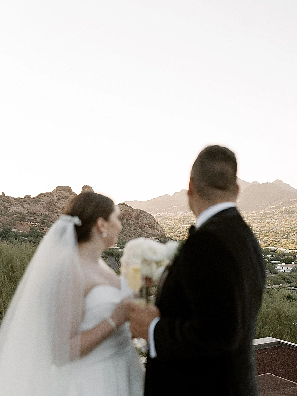 Couple portrait of bride and groom from behind toasting champagne, her veil and bouquet visible on a rooftop terrace over desert mountains.