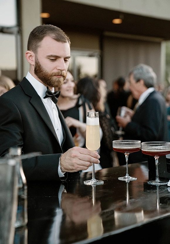 Groom portrait at bar in a black tuxedo and bow tie, holding a champagne flute by coupe cocktails on an evening-lit patio with guests