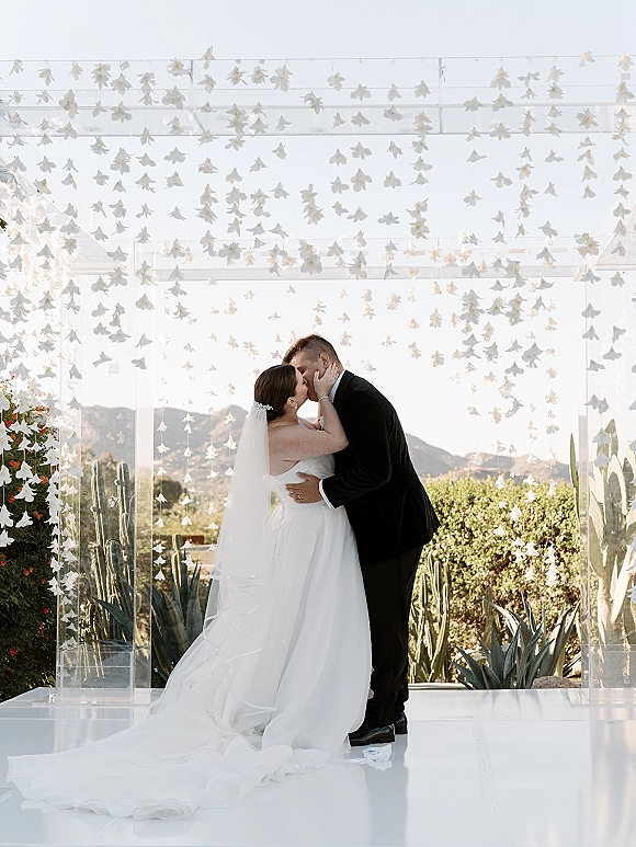 Wedding kiss portrait of bride and groom kiss under a clear acrylic arch with hanging white flowers, desert cactus and mountains behind