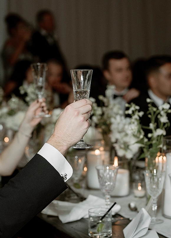 Wedding toast with champagne toast wedding flutes raised over a candlelit reception table with white florals, crystal glassware, and tuxedo cufflinks
