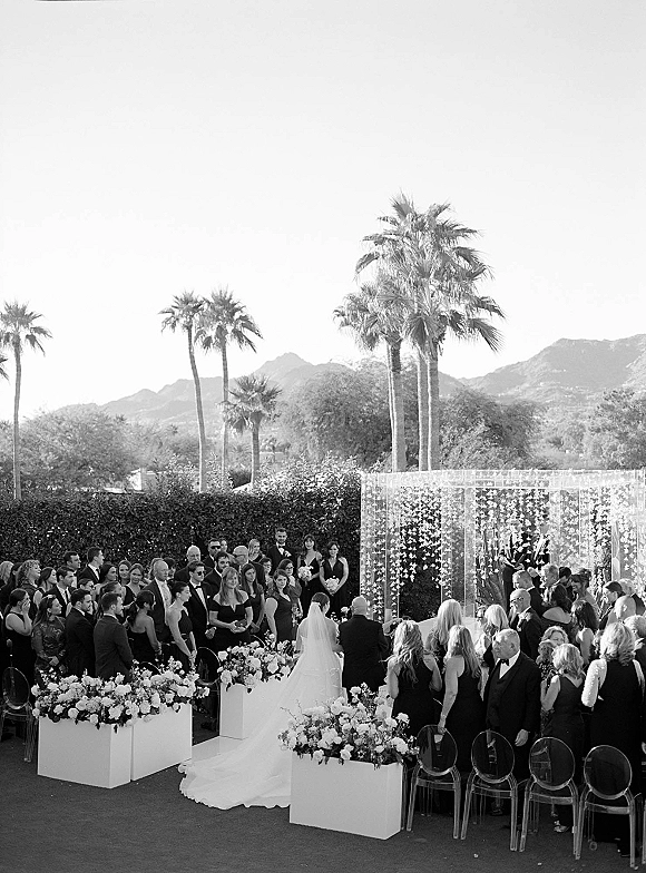 Wedding ceremony outdoors with bride walking down a white aisle past large florals and clear chairs toward an arch, palm trees and mountains behind
