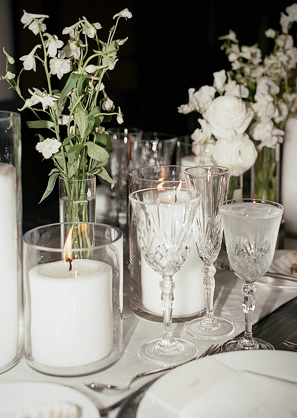 Reception tablescape with a wedding table centerpiece of white florals in bud vases, greenery and pillar candles in glass against dark drapery