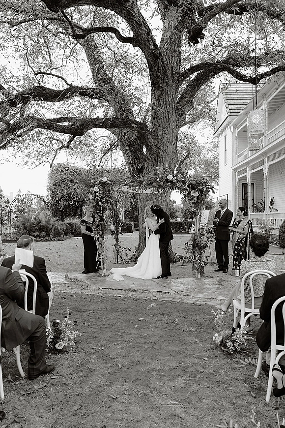 Ceremony kiss moment at an outdoor wedding ceremony, bride and groom under a floral arch beneath a large tree on a lawn near a house porch