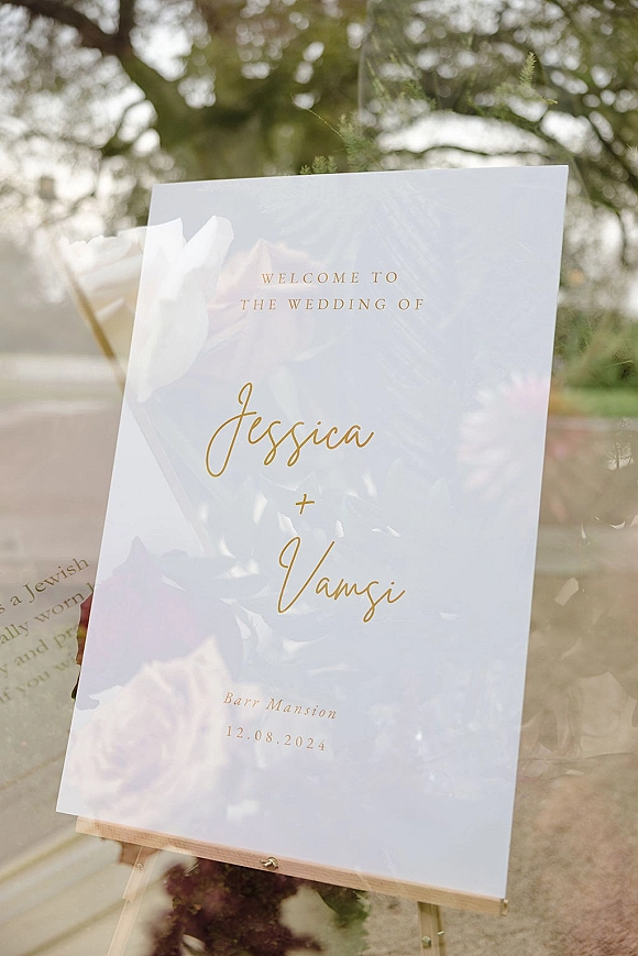 Wedding welcome sign acrylic with gold lettering on an easel, set on a lawn with trees and outdoor greenery in the background