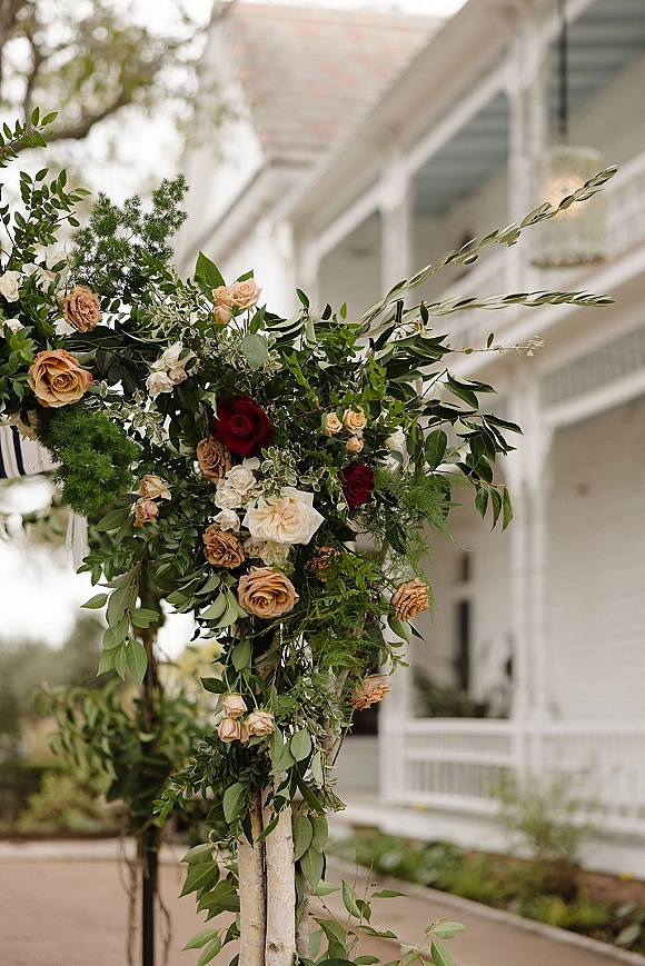 Wedding ceremony florals with roses and greenery arranged asymmetrically on birch poles by a porch railing with trees beyond