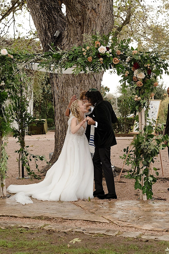 Wedding ceremony moment as bride and groom kiss under a floral chuppah with greenery garland and roses in an outdoor garden setting