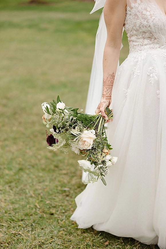 Bridal portrait of a bride holding bouquet with a white-and-green wedding bouquet, lace dress and veil, henna hands on a grass lawn