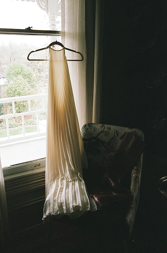 Wedding dress on hanger, a pleated bridal gown hanging by a window on a porch with railing and trees beyond, softly lit by daylight