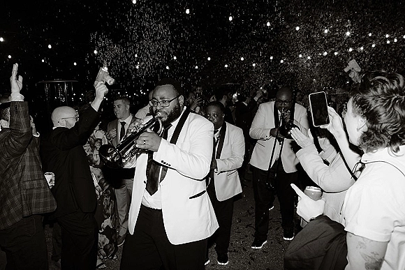 Wedding band leading a brass band wedding entrance as guests cheer and film under string lights, confetti falling on a night street