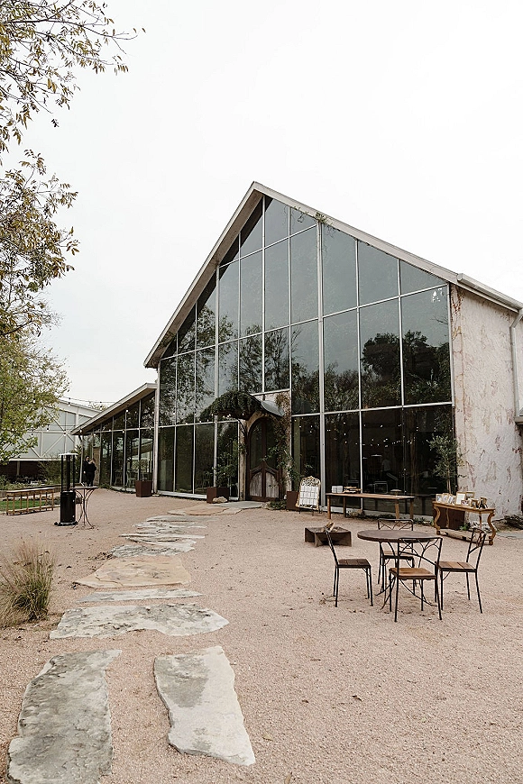 Wedding venue exterior with string lights over patio tables and metal chairs, leading to wooden doors of a glass greenhouse in a gravel courtyard