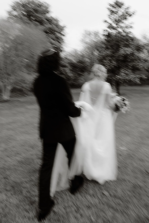 Wedding couple portrait in black and white, bride and groom walking away on a grassy lawn, veil trailing and bouquet in hand