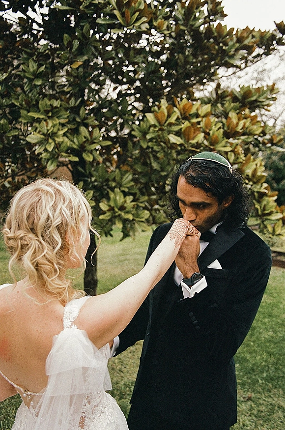 Ceremony moment as groom kissing bride hand, her henna and lace bodice visible, while they hold hands on a grassy lawn by a tree