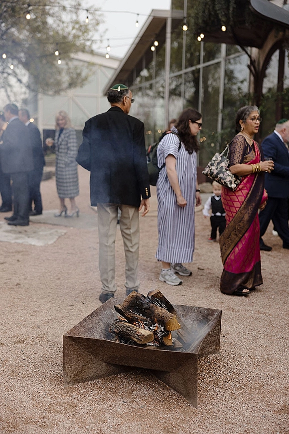 Wedding cocktail hour guests mingling at an outdoor cocktail hour around a metal fire pit with string lights, drinks, suits, sari, and kippah in a courtyard