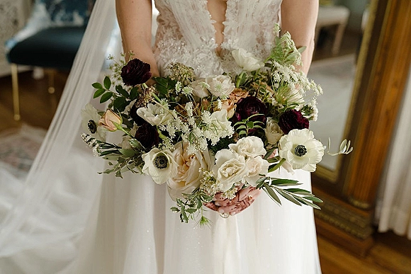 Bridal bouquet of white and burgundy flowers with blush roses and greenery held against a lace bodice, with veil by a mirror indoors