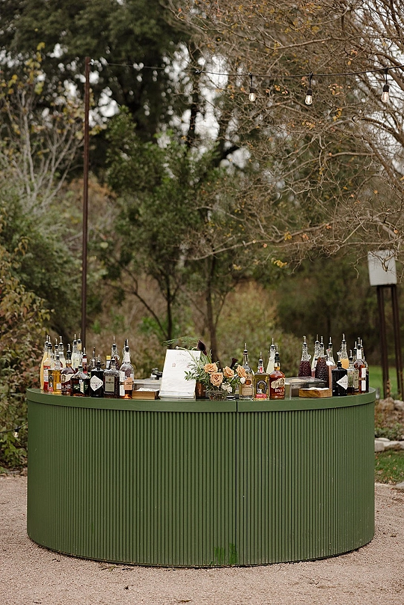 Wedding bar setup with a curved bar, liquor bottles and glassware, plus a floral arrangement and menu sign under string lights in a garden setting