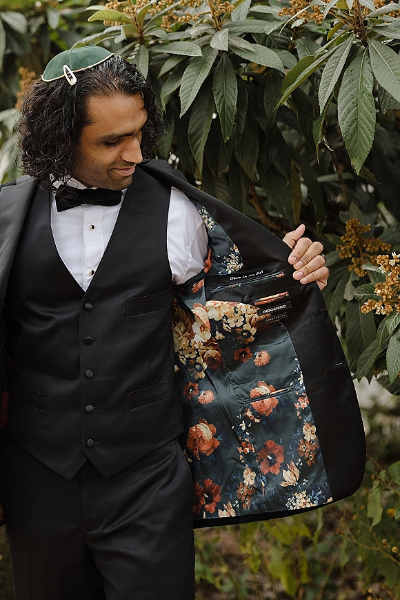Groom portrait in a black tuxedo and waistcoat with bow tie, showing floral jacket lining, standing before lush garden greenery