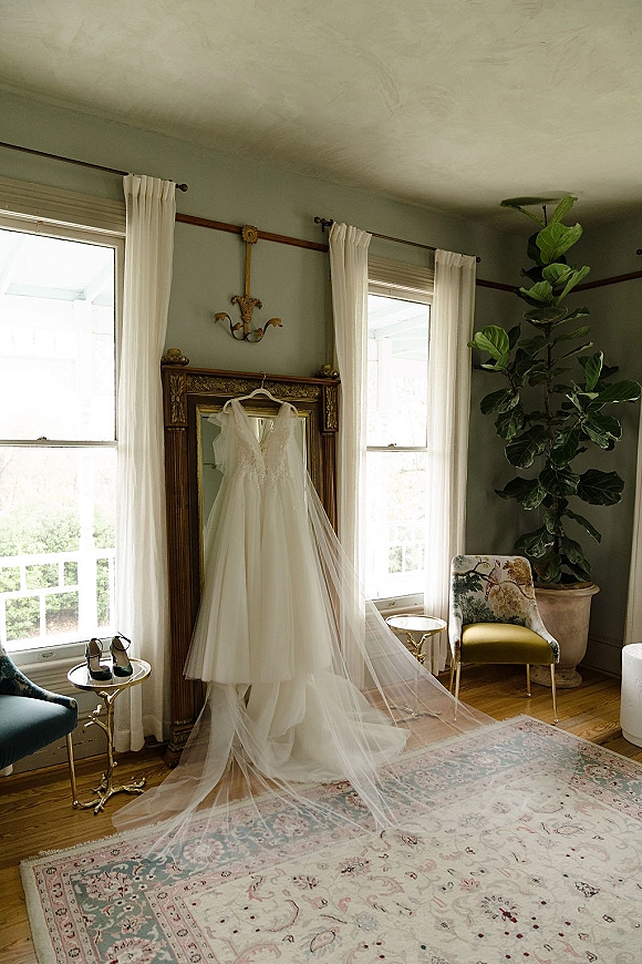 Wedding dress hanging by sunlit windows with long veil and heels nearby, reflected in an antique mirror on wood floors