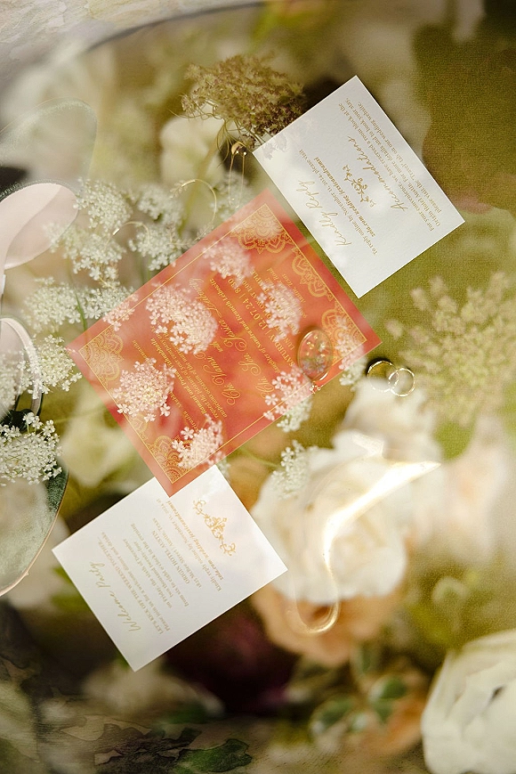Invitation suite in a wedding invitation flat lay with red card and gold foil lettering, rings, white flowers and ribbon on greenery backdrop