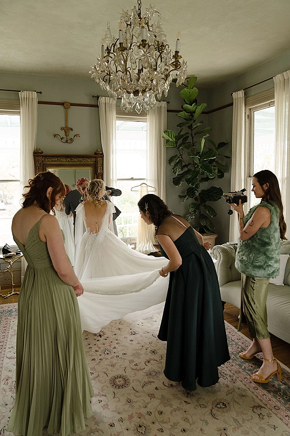 Bride getting ready as bridesmaids fluff bridal dress train and veil in a bright living room with large windows and a vintage chandelier