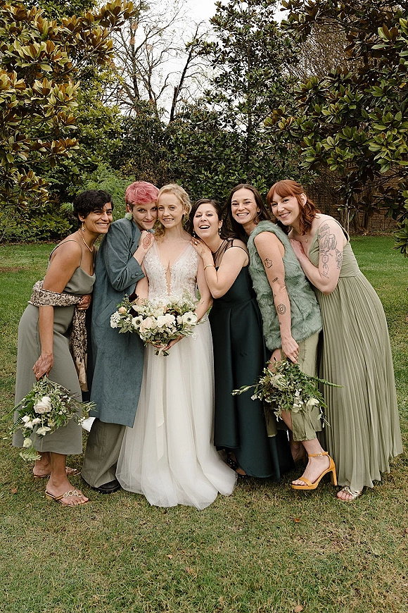 Bridesmaid group photo with bride with bridesmaids holding white and greenery bouquets on a lawn with garden trees under an overcast sky