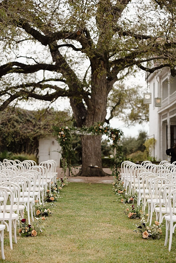 Ceremony setup for an outdoor wedding ceremony with white chairs and a rose-lined aisle leading to a floral arch on a lawn by a white house