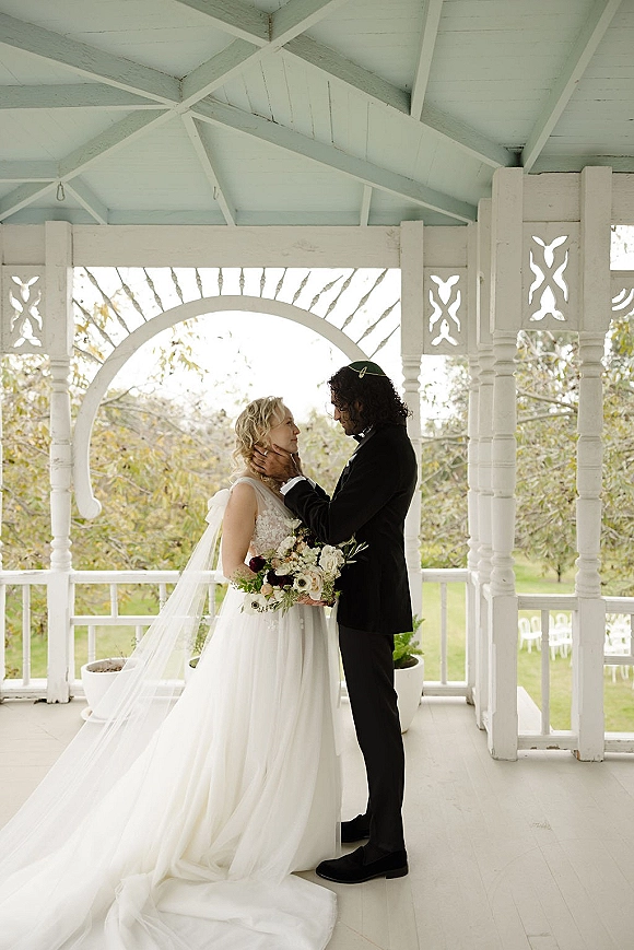 Couple portrait of bride and groom embrace as he holds her face, lace dress and veil beside a white gazebo porch with trees beyond