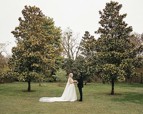 Couple portrait of bride and groom holding hands, facing each other on a lawn with trees, her veil and long train in soft light