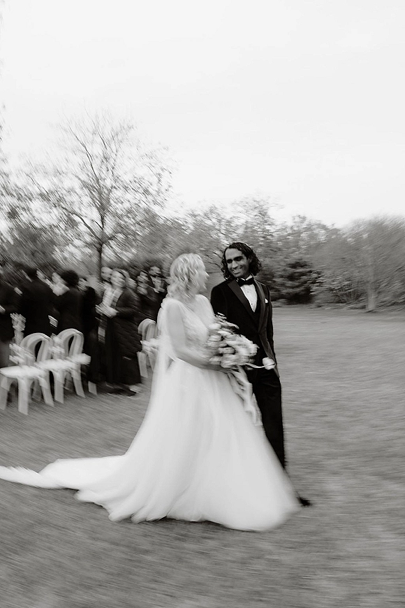 Wedding recessional as bride and groom walking down the aisle, she holds a bouquet while guests cheer on a grassy lawn with trees