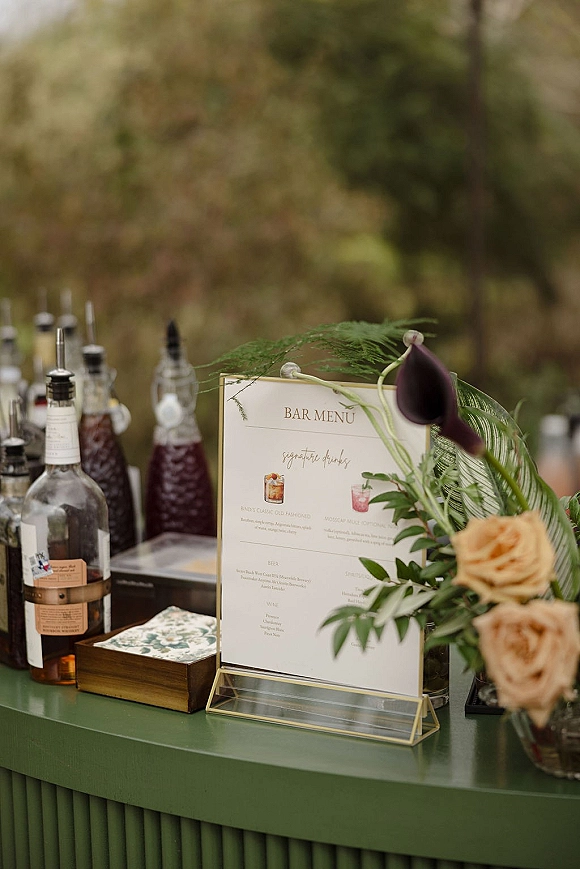 Wedding bar menu in an acrylic holder beside liquor bottles and a floral arrangement with a calla lily, set against blurred outdoor greenery