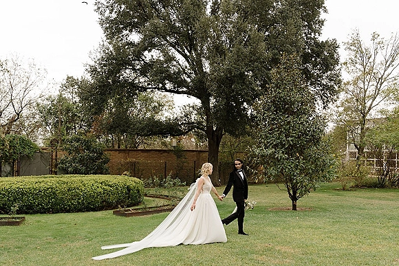 Couple portrait of bride and groom holding hands, her long veil and bouquet trailing as they walk on a garden lawn by a brick wall gate