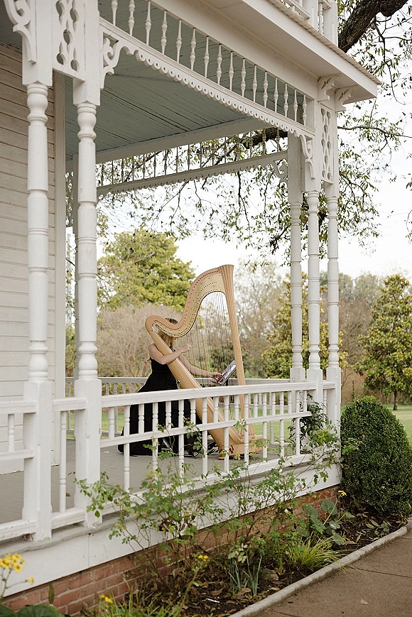 Wedding harpist in a black dress playing a concert harp on a white wraparound farmhouse porch with railings and trees beyond