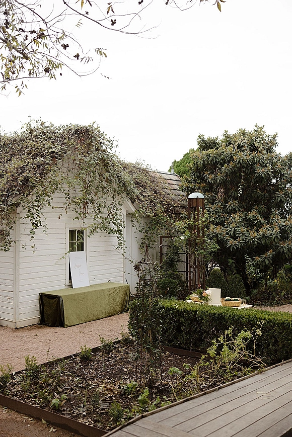 Wedding welcome table with welcome table decor, a sign on an easel, baskets, and flowers set by an ivy-covered white cottage in a garden