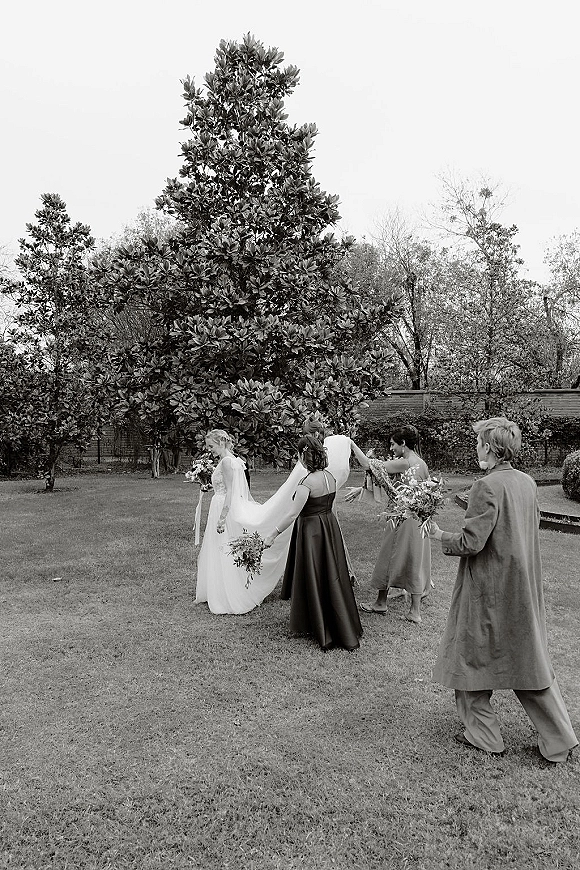 Getting ready moment as bridesmaids help the bride adjust her bridal veil, holding bouquets on a garden lawn by a brick wall
